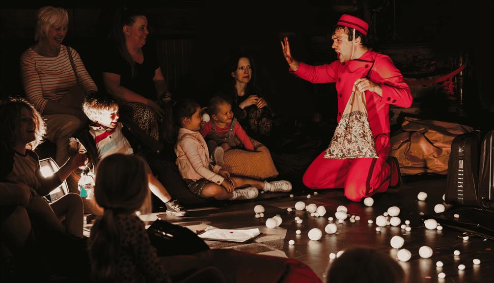 Actor on stage dressed in red with audience of adults and children watching