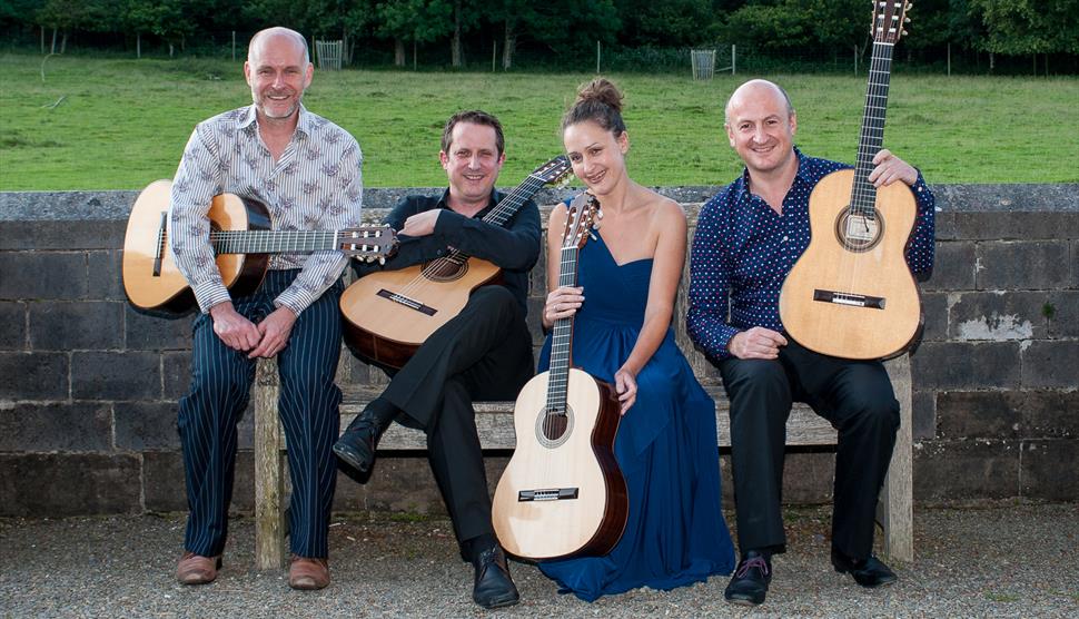 Three men and one woman sit on a bench in front of a field. They are all holding guitars.