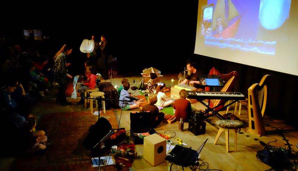 A group of children are sat in front of a movie screen with instruments like shakers, cymbals, recorders, guiros, a gong and xylophone..