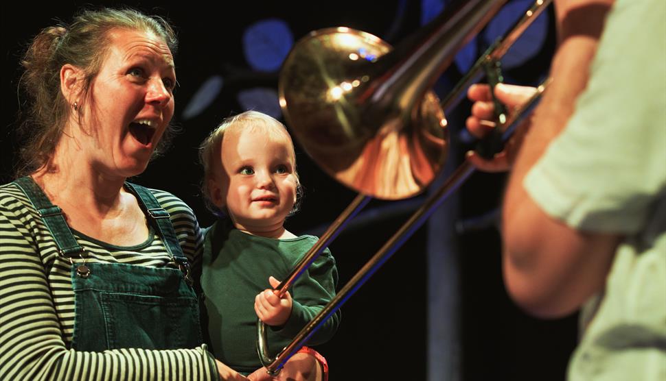 A mother holds her toddler. The toddler is grabbing at a trombone slide from a trombone player off screen.