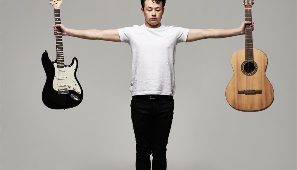 Sean Shibe stands holding two guitars, an electric guitar to the left and an acoustic guitar to the right
