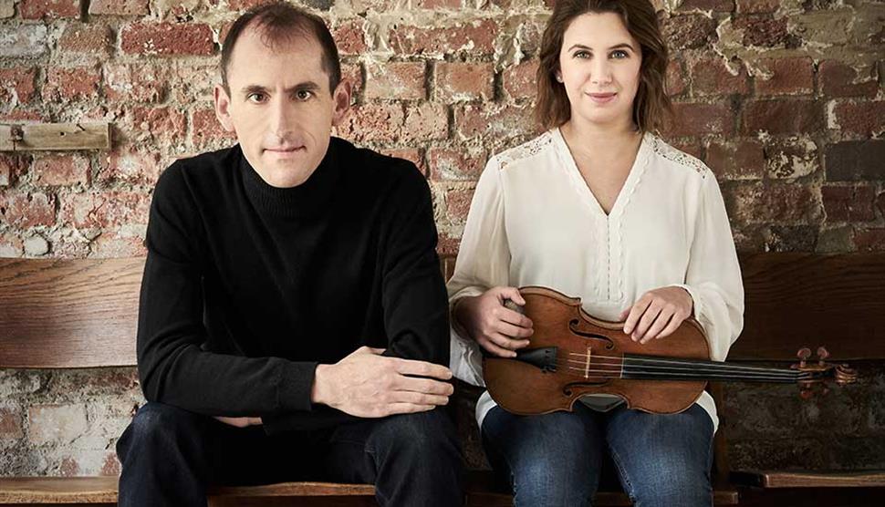 Chloë Hanslip and Danny Driver sit next to each other in front of a brick wall looking at the camera.