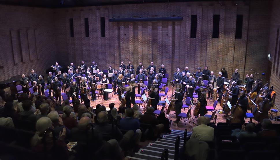 The image has a full orchestra standing on Turner Sims' stage. The orchestral players and conductor are looking towards the audience.