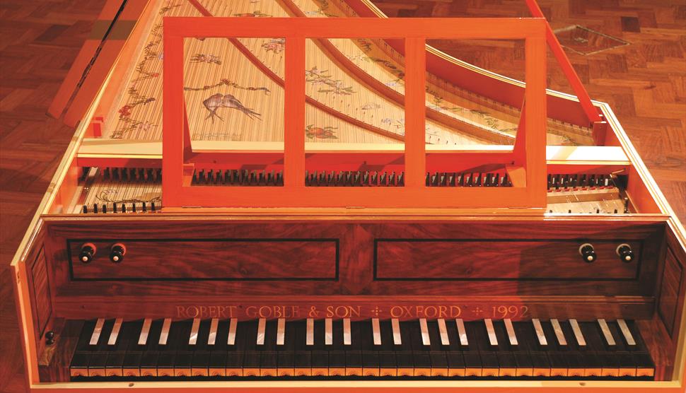 An old harpsichord made of wood, with etchings of birds and flowers on the top. Above the keys, etched are the words 'Robert Goble & Son / Oxford / 19
