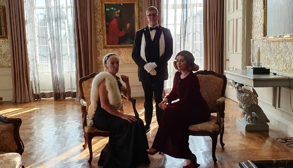 A man stood up and two women sat in chairs dressed in 1920s clothing in a opulent room.