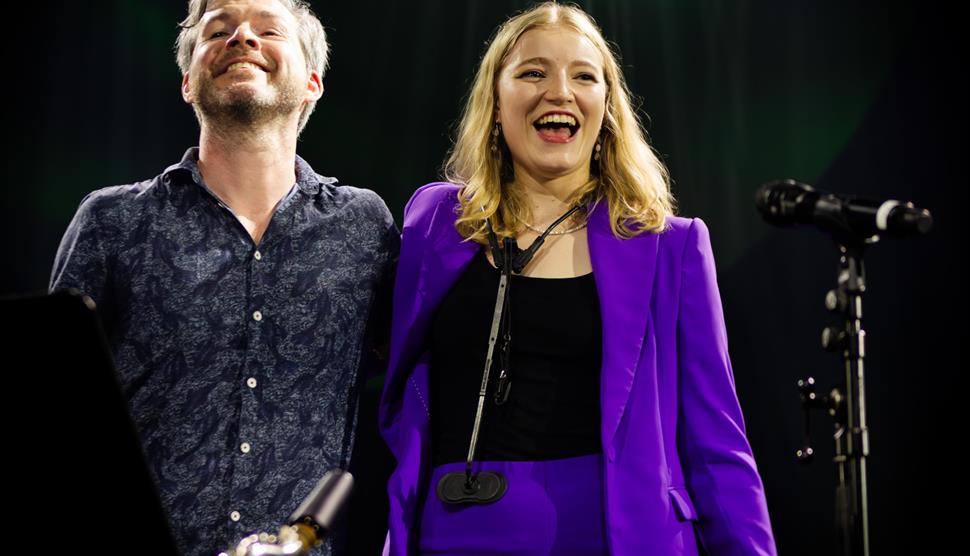 Emma Rawicz and Gwilym Simcock stand side by side smiling next to a microphone.