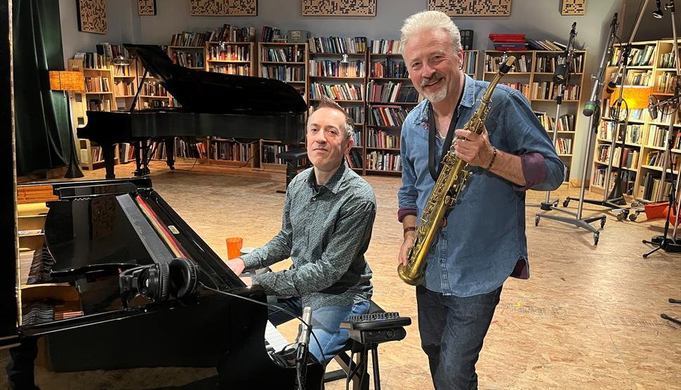 One man sits at a piano while another man stands next to him, holding a mezzo-soprano saxophone and smiling.