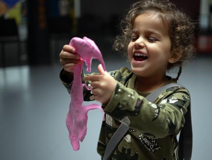 A young girl holding pink slime. The girl is looking at the slime with a big smile on her face.