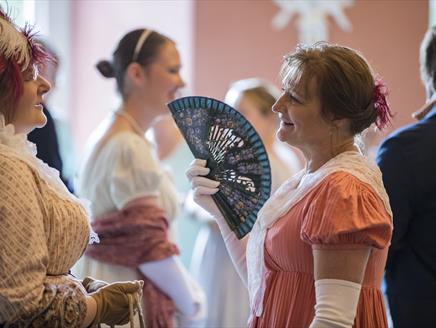 Ladies in Regency costume chat at an event. A lady on the right uses a black fan.