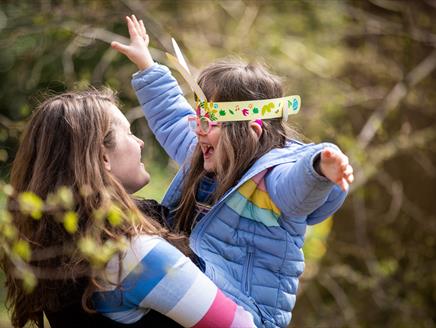 A parent lifts their child up. The child wears bunny ears and a lilac coat and has their arms up in the air.