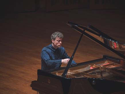 Paul Lewis is playing the piano in a dimly lit concert hall.