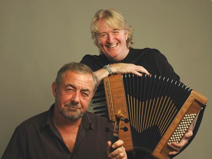 Phil Cunningham is standing, holding his accordion and smiling, with Aly Bain sitting in front of him holding his fiddle.