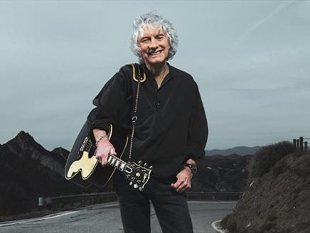 Albert Lee wearing a black shirt and smiling at the camera, holding a guitar and standing on a road with mountainous terrain in the background