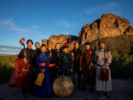 A group of musicians wearing traditional Mongolian clothes and stand in front of a hill. They're all holding traditional Mongolian instruments.