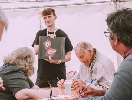 Poet delivering workshop, holding up a book, with participants sitting at a table.