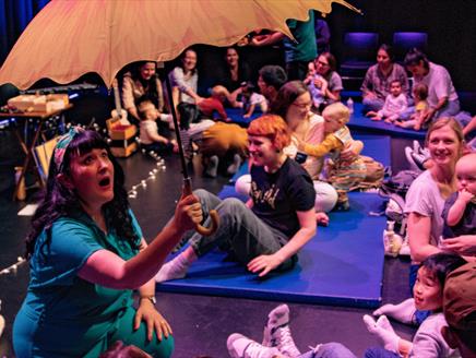 A woman with black hair is sat down and holding a yellow umbrella. Parents and babies are looking at her while sat on the floor.