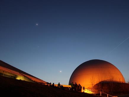 A dark sky with a few stars. The Science Centre and Planetarium are glowing orange against the night sky.