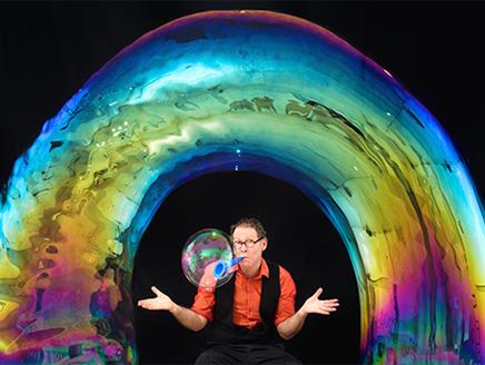Louis Pearl (The Amazing Bubble Man) sitting underneath a bubble in the shape of a rainbow, with his hands outstretched. He is also blowing a bubble f