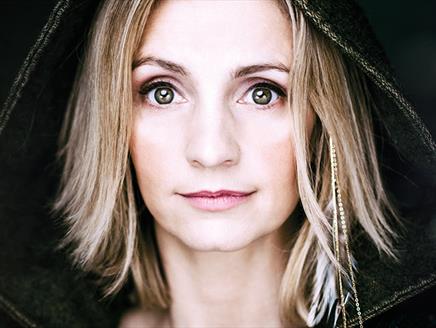 A blonde woman wearing a black hood looks towards the camera.