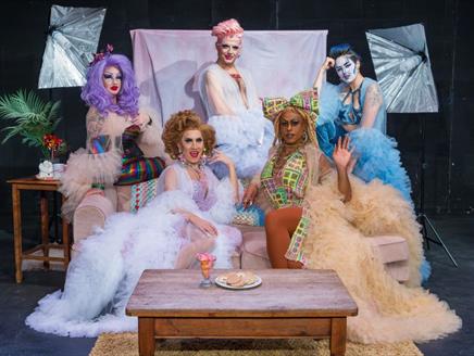A group of drag queens and cabaret artists in colorful costumes poses together on a couch, smiling and enjoying their time.