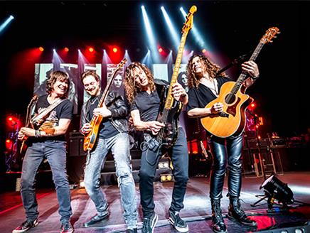Four men playing guitar on a stage with strobe lighting in the background