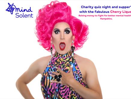 Drag artist Cherry Liquor is pulling a shocked face with their mouth open. They're wearing a big pink styled wig, dangling jewelled earrings and a mul