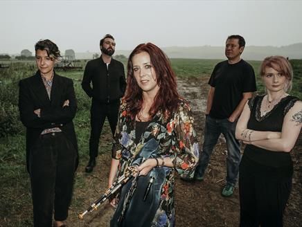 Five people in dark clothing stand on a muddy path in the countryside. A woman with red hair and a dark floral jacket stands in the centre.