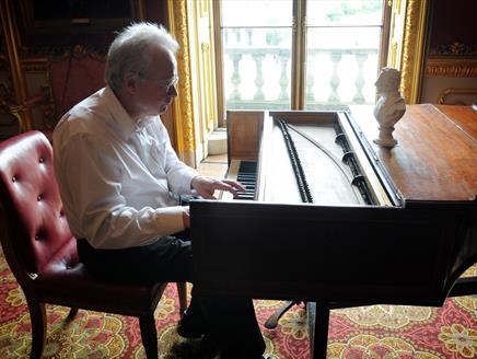 The image shows David Owen Norris sat on a red chair playing a piano by the window.