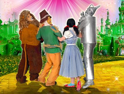 Four actors dressed as the Cowardly Lion, Scarecrow, Dorothy, and the Tin Man from The Wizard of Oz. They are all linking arms with the Cowardly Lion,