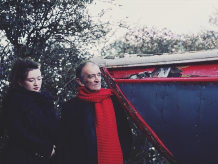 A woman with dark hair and a black coat stands next to an older man wearing a red scarf.