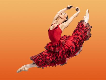Ballet dancer in red dress leaps through air. Orange background.