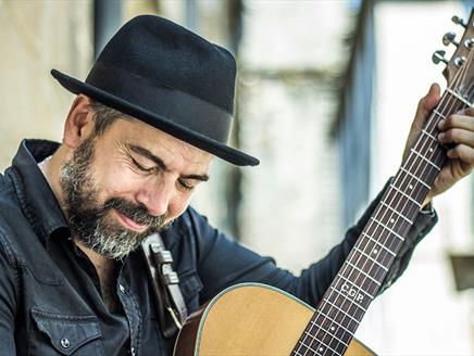 Iago Bennet, a man with a short greying beard, wearing a fedora, and dark blue button up shirt, playing the guitar with a very content look