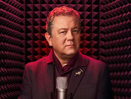 Jon Culshaw wearing a dark suit with a patterned shirt and a pin on his lapel. He's standing in front of a professional microphone soundproofing foam