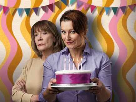 Two actresses stood in front of a 70s style wall, it has a wavy pattern with cream, yellow, brown, and light pink colours. There is multi coloured bun