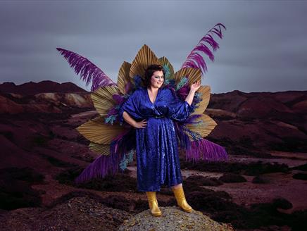 Kiri Pritchard-McLean stands in a sparkly blue dress on a rock, with a backdrop of a gloomy moor. Attached to her back is a large display of feathers,