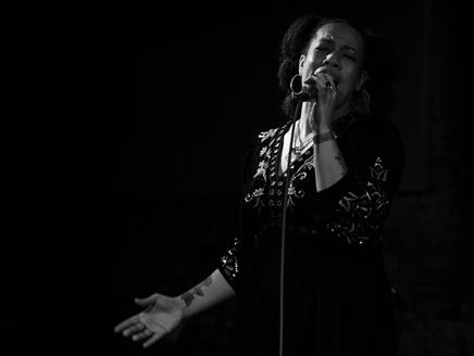 A black-and-white image of a woman singing into a microphone. She has large hoop earrings and a black top with embroidered leaves.