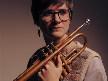 A woman with short hair glasses holds a trumpet.