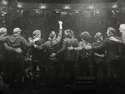 Black and white photo of the backs of nine members of the Levellers on stage, looking towards the audience with their arms around each other