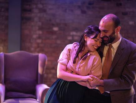 Two actors on stage, man is holding woman in an embrace. They are both smiling, they look in love. There is an empty chair behind them