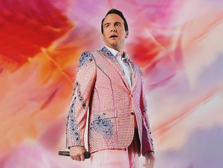 A man in a sparkly pink suit - Matt Forde - holds a microphone, standing against a vibrant, swirling pink and red background.