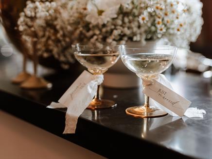 2 champagne coupe glasses sits on a bar labelled Mr & Mrs in front of wedding flowers and a discoball.