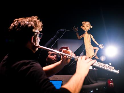 A flute player faces away from the camera whilst playing their instrument in front of a puppet being controlled by a puppeteer.