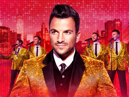 Peter Andre wearing a gold sparkly jacket stood in front of four other male singers wearing the same outfit, singing into microphones, on a red city s