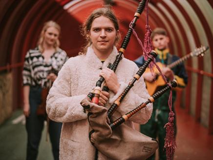 A man with long, brown hair and a white coat carries a bagpipe.