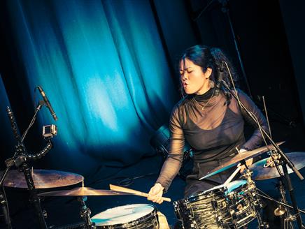 Sun-Mi Hong is sat in front of blue curtains and behind a drum kit and appears to be passionately drumming with her eyes closed.