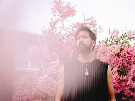 A man with dark hair and a black jumper stands in front of a cherry blossom tree.