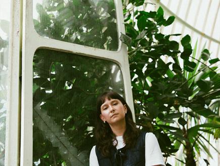 A woman with dark brown hair stands in front of dark green leaves in a forest.