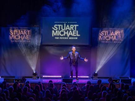 Stuart Michael stands on stage in front of an audience, there are three screens behind him with his name on.
