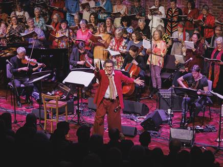 Harvey Brough reaches his arm out and smiles towards the camera. A choir and multiple instrumentalists are behind him.