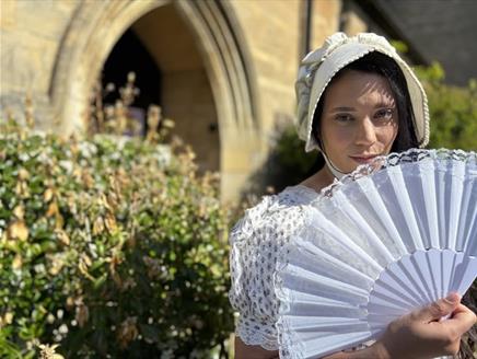 a woman dressed in white holding a fan standing in front of bushes